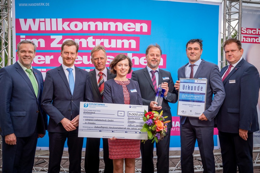 Männer und Frauen stehen vor einer Plakatwand. Eine Frau hält einen Scheck in der Hand.