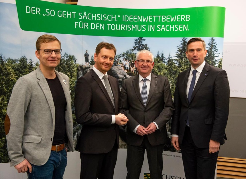 Vier Männer stehen vor einer touristischen Plakatwand, die das Elbsandsteingebirge zeigt.