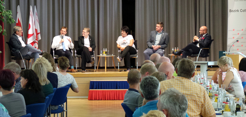 Ein Saal in dem Menschen versammelt sind und Männern und Frauen auf einem Podium zuhören.