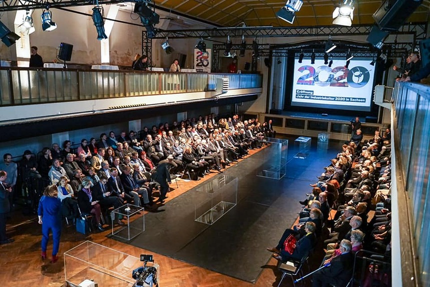 In einer großen Halle sitzen Menschen vor einer Bühne, auf der Glasvitrinen stehen.