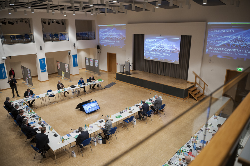 Männer und Frauen sitzen in einer Halle an einem Konferenztisch.