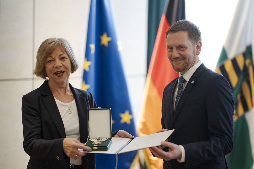 Ein Mann überreicht einer Frau einen Orden. Im Hintergrund steht eine Fahnengalerie.