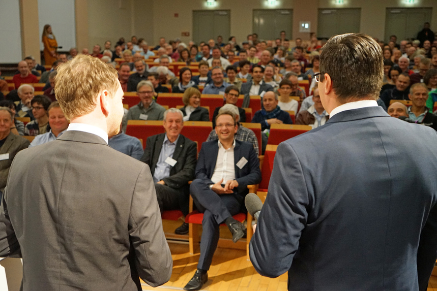 Zwei Männer stehen auf einer Bühne vor einem versammelten Publikum.