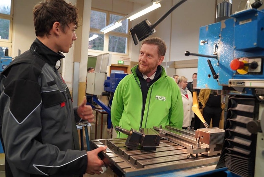Ein Mann in einer grünen Jacke unterhält sich mit einem jungen Mann im Arbeitsanzug an einer Maschine.