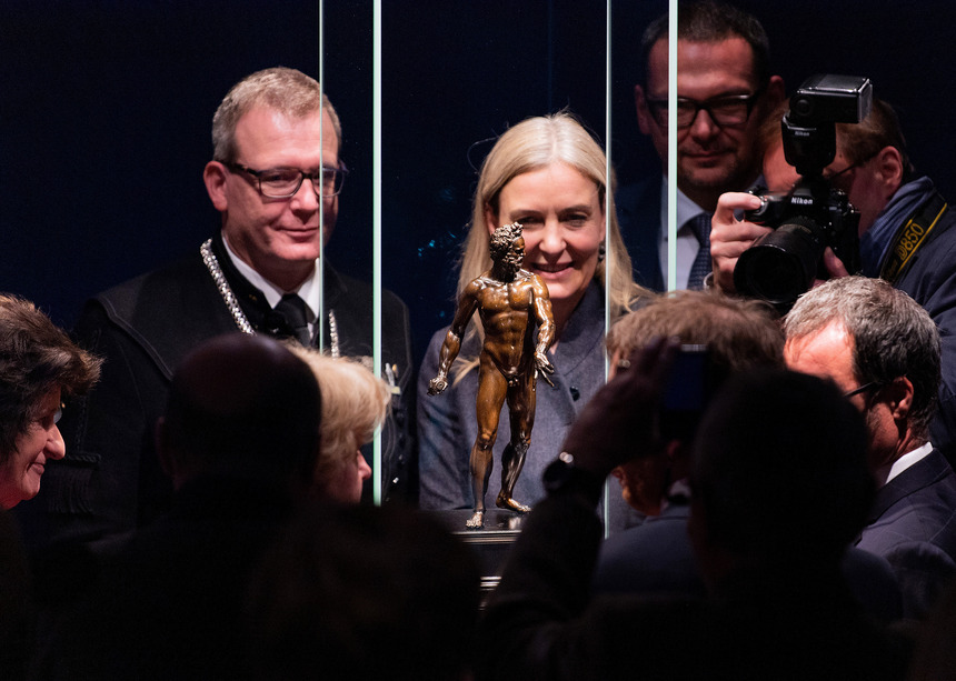 Männer und Frauen stehen vor einer Glasvitrine, in der eine Bronzestatue eines Mannes ausgestellt wird.