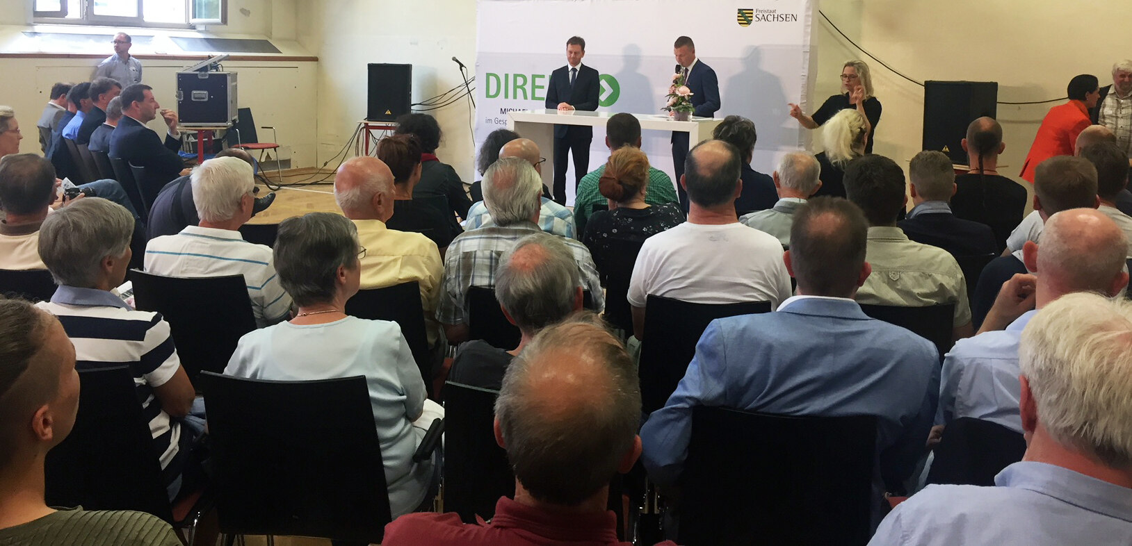 Zwei Männer stehen vor einer Plakatwand in einer Turnhalle. Vor Ihnen sitzen Männer und Frauen.