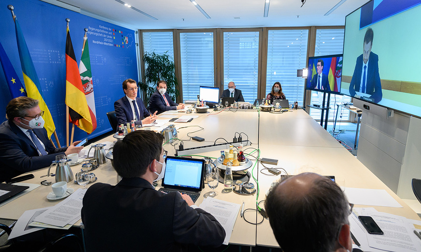 Männer und Frauen sitzen an einem Konferenztisch und blicken auf einen Monitor. Im Hintergrund steht eine Fahnengalerie.