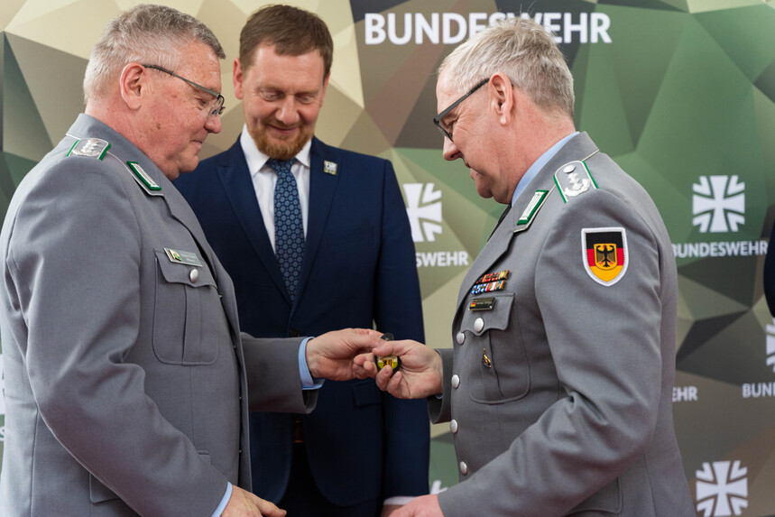 Drei Männer stehen sich gegenüber. Zwei Männer reichen sich eine Marke in die Hand.