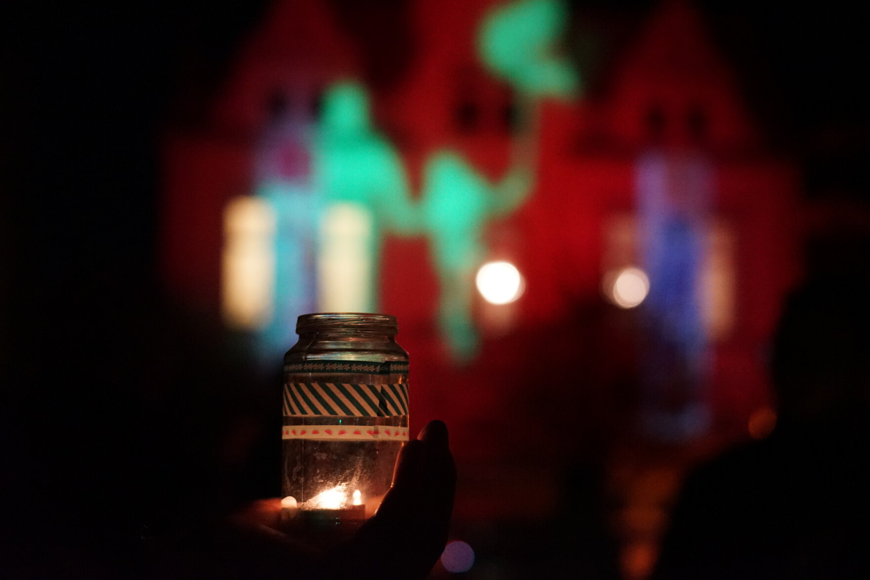 Eine Perosn hält ein Teelicht. Im Hintergrund sind Lichtspiele an einer Hausfassade zu sehen.