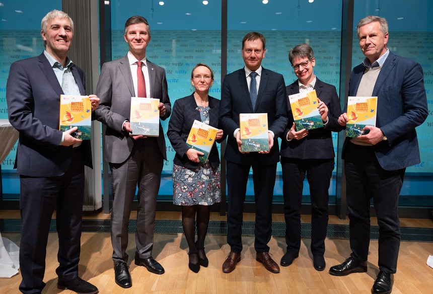 Männer und Frauen stehen auf einer Bühnen und halten ein Chorbuch in der Hand.