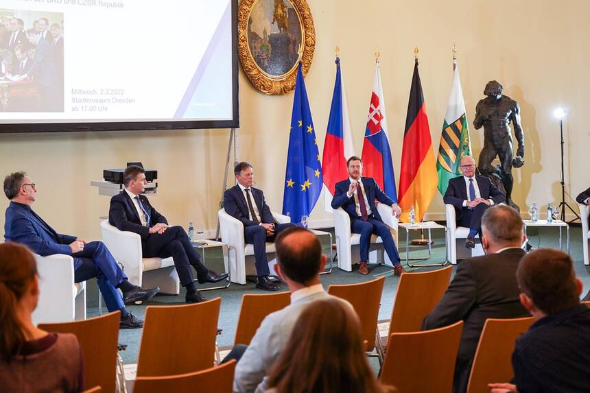 Männer sitzen auf einem Podium vor einer Leinwand. Neben ihnen steht eine Fahnengalerie.