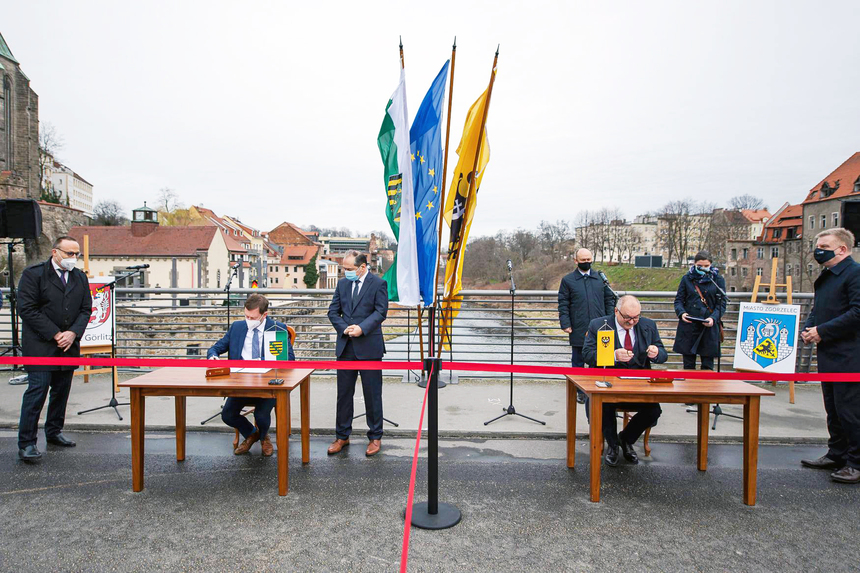 Männer sitzen an Tischen auf einer Brücke und unterzeichnen ein Schriftstück.