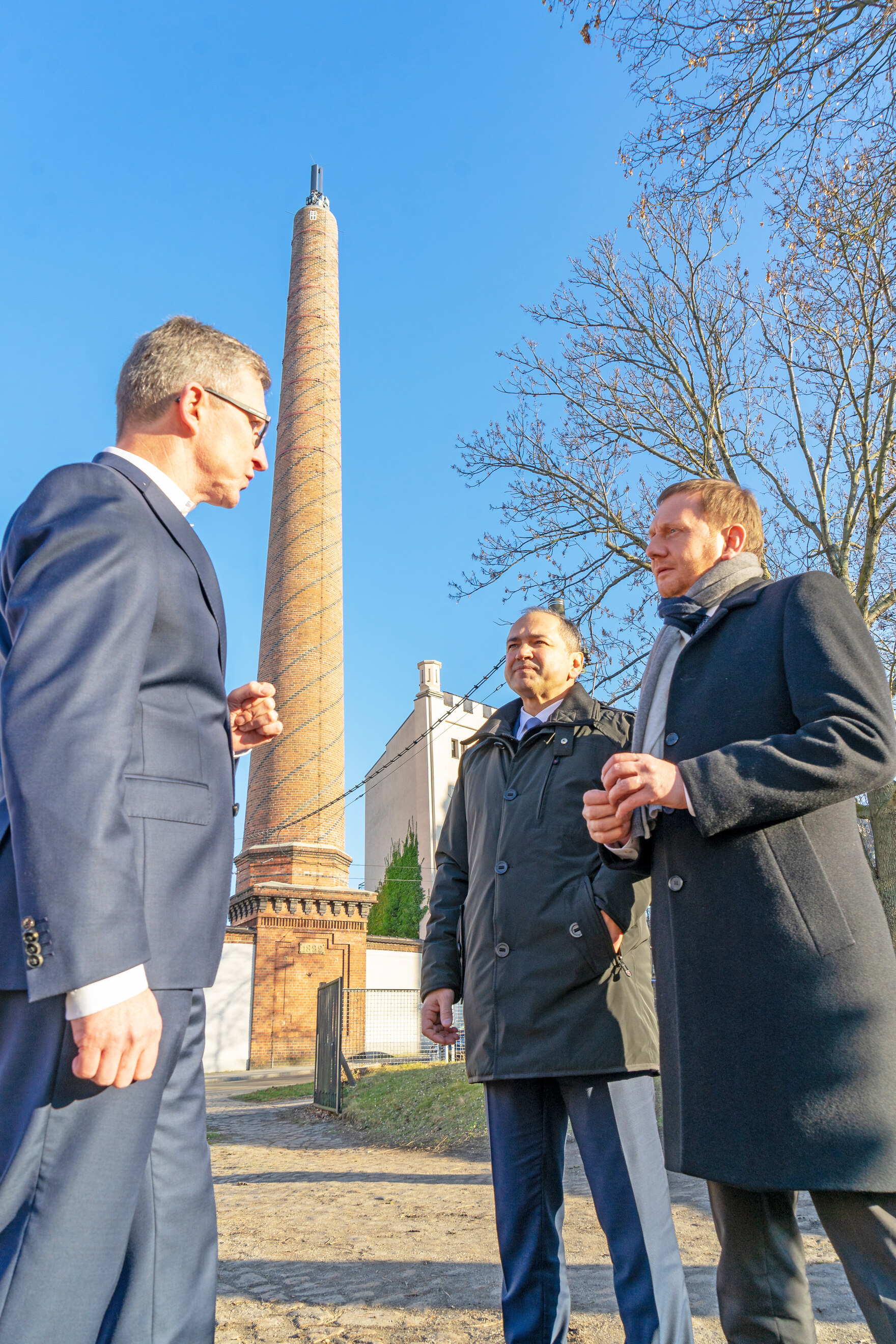 Drei Männer stehen vor einem alten Schornstein, auf dem eine Mobilfunksendemastanlange steht.