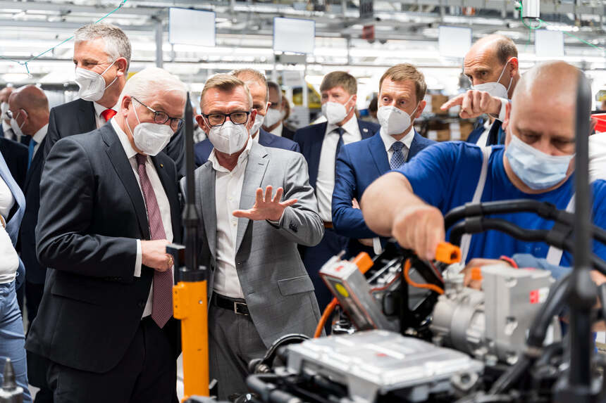 Männer stehen in einem Automobilwerk und schauen einem Fließbandmitarbeiter bei der Arbeit zu.