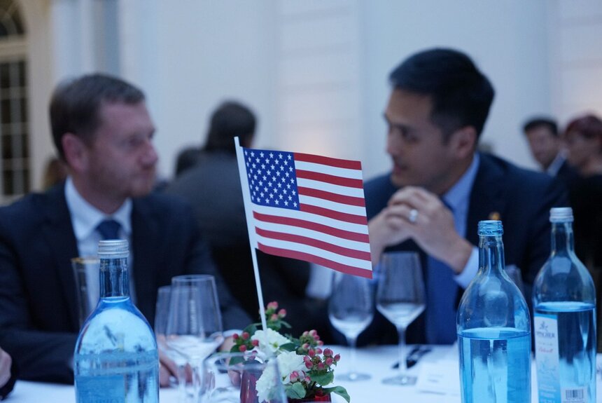 Eine amerikanische Flagge steht auf einem Banketttisch. Im Hintergrung sitzen zwei Männer und unterhalten sich.