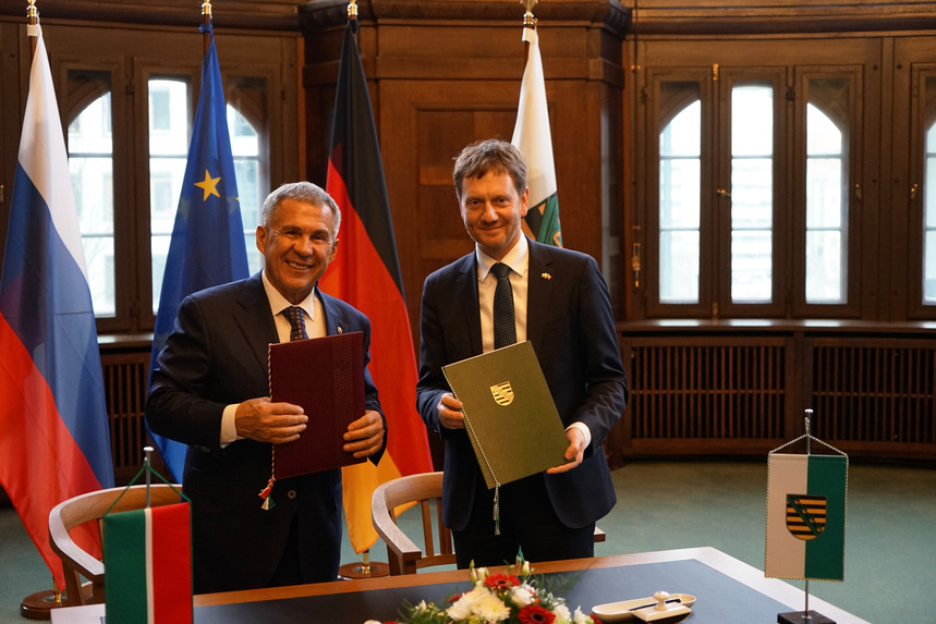 Zwei Männer stehen vor einem Tisch und halten jeweils eine Mappe in der Hand. Im Hintergrund steht eine Fahnengalerie.