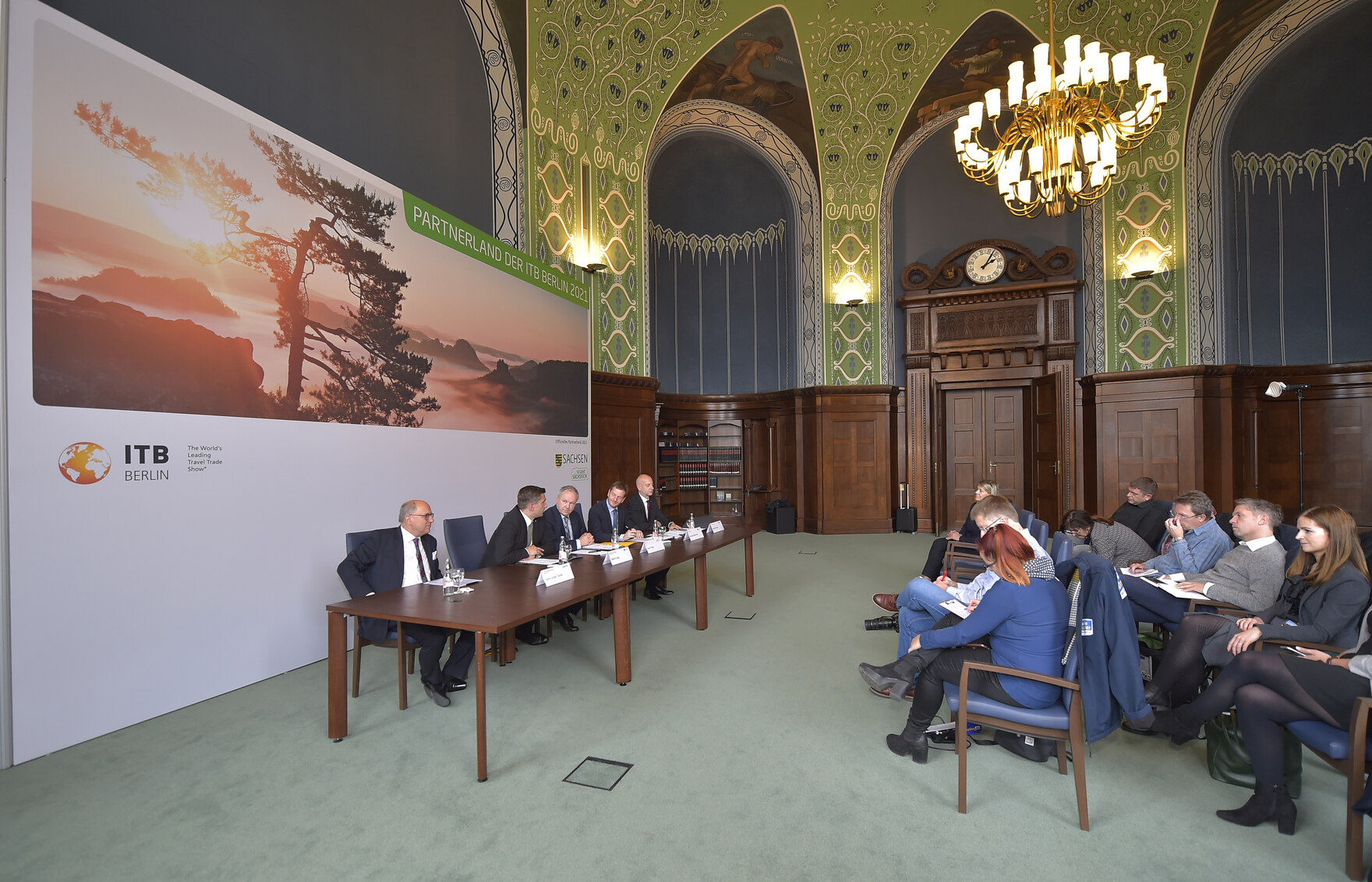 Männer sitzen vor einer Leinwand in einem großen Saal. Davor sitzen Journalisten.