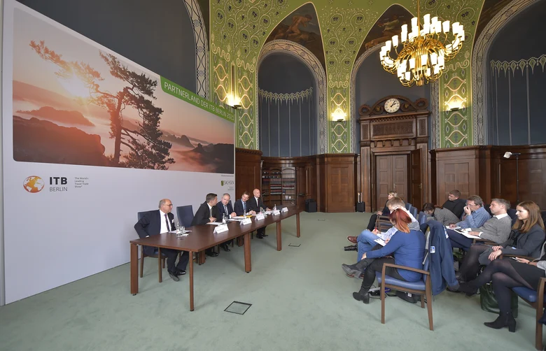 Männer sitzen vor einer Leinwand in einem großen Saal. Davor sitzen Journalisten.