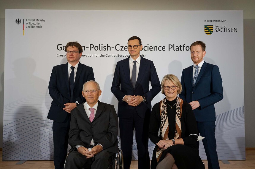 Männer und eine Frau posieren vor einer Pressewand für ein Foto.