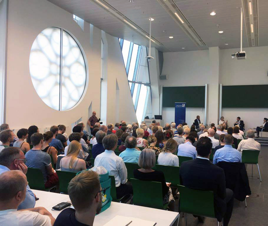 Menschen sitzen in einem Hörsaal und beobachten Podiumsteilnehmer am Ende des Saals.