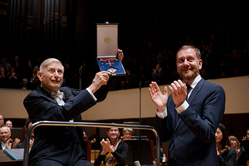 Zwei Männer stehen vor einem Orchester. Einer hält einen Verdienstorden in der Hand und hebt ihn in die Höhe.