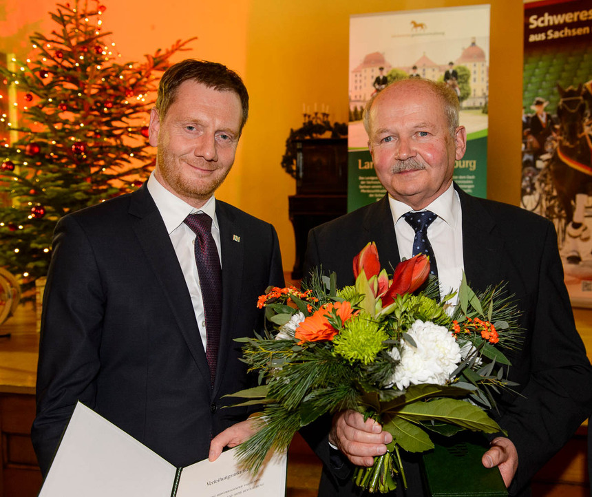 Zwei Männer stehen vor einer Bühne. Einer von ihnen hält eine Urkunde in der Hand, der andere einen Blumenstrauß.