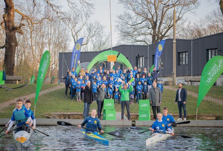 Männer und Frauen stehen am Ufer. Vor Ihnen fahren Kanuten im Wasser.