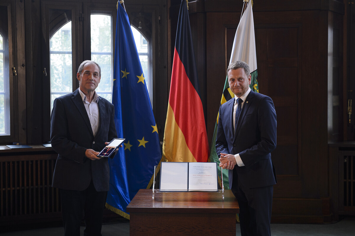 Zwei Männer stehen an einem Pult. Einer von beiden hält einen Orden in der Hand.