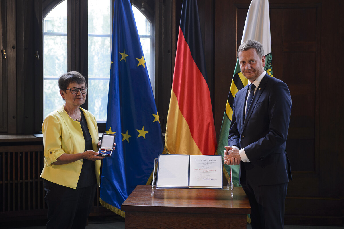 Eine Frau und ein Mann stehen neben einem Pult. Die Frau hält einen Orden in der Hand.