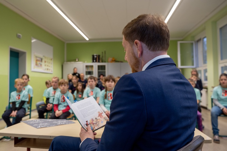 Ein Mann sitzt vor einer Gruppe Schülerinnen und Schüler und liest ihnen aus einem Buch vor.