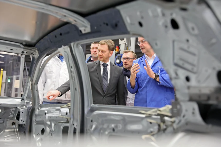 Ein Auto im Rohzustand auf einem Förderband, dahinter stehen zwei Herren.