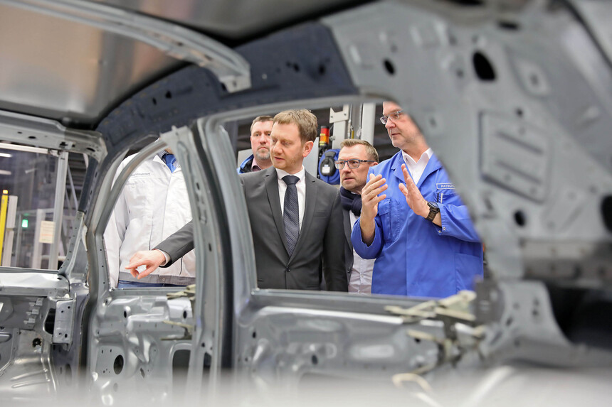 Ein Auto im Rohzustand auf einem Förderband, dahinter stehen zwei Herren.