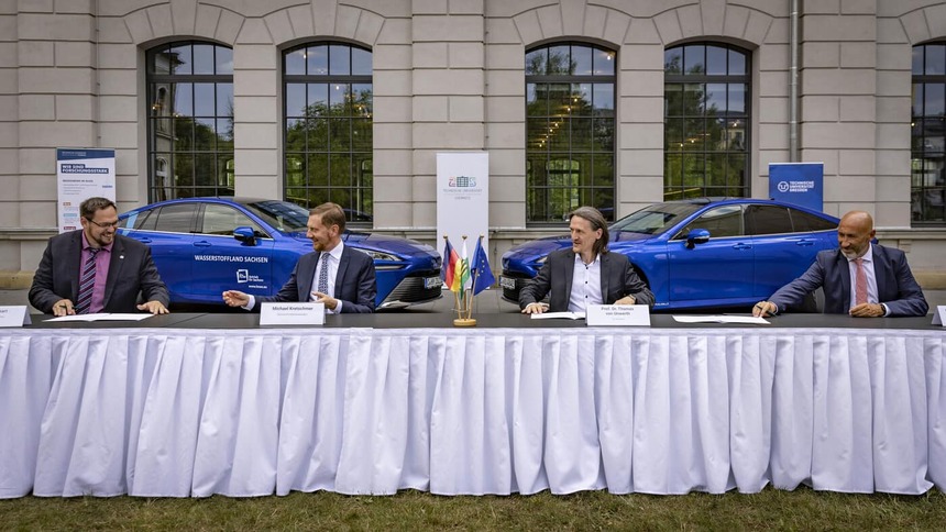Männer sitzen an einer Tafel. Im Hintergrund stehen zwei Autos.