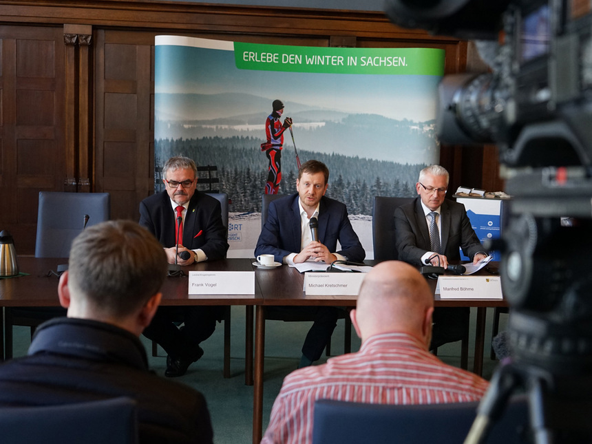 Männer sitzen vor einer Pressewand an einem Tisch und sprechen mit Journalisten.