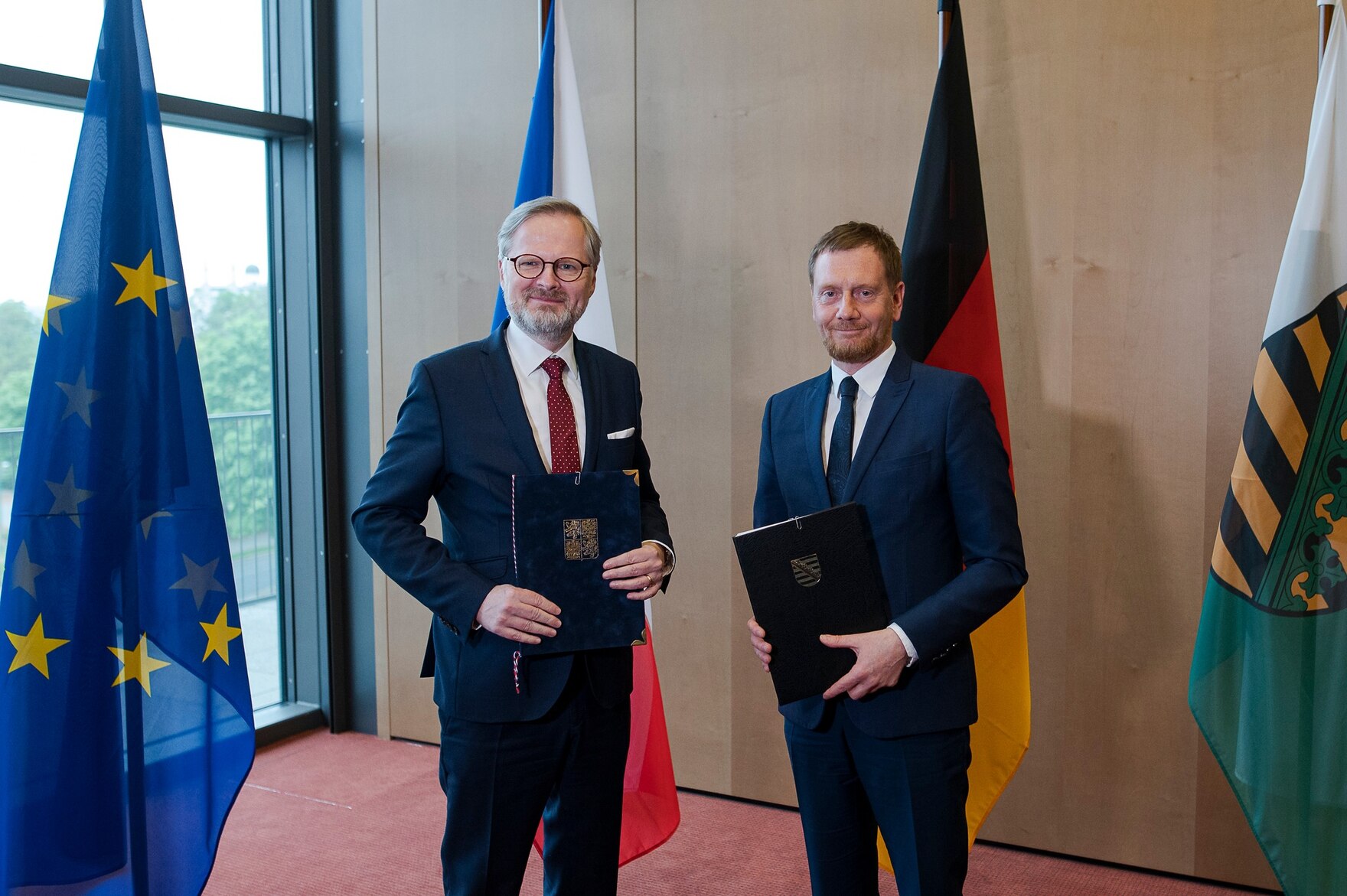 Zwei Männer stehen vor einer Fahnengalerie und halten eine Mappe in der Hand.