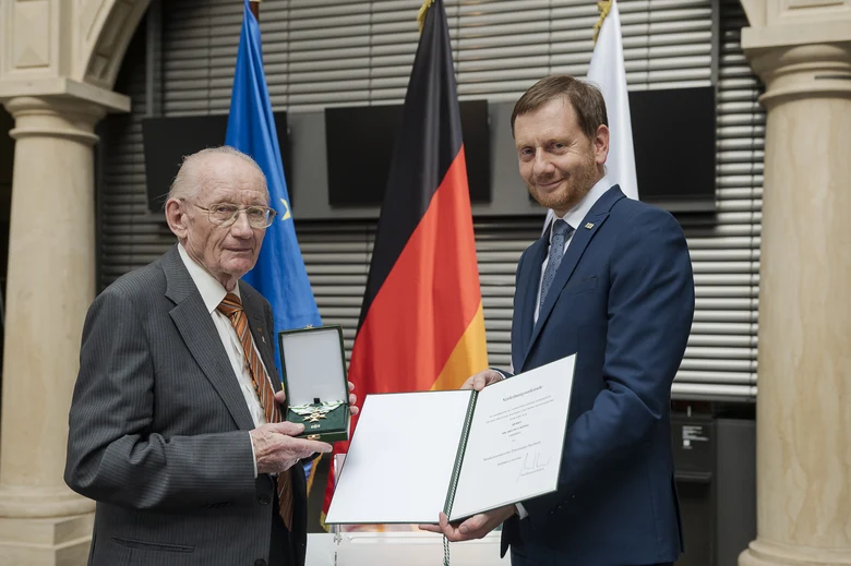 Ein Man hätl eine Urkunde in der Hand. Neben ihm steht ein weiterer Mann.
