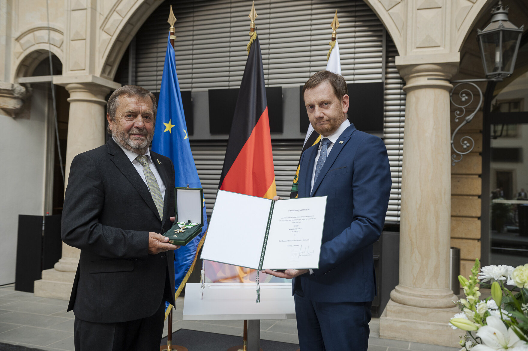 Ein Mann hält eine Urkunde in der Hand. Neben ihm steht ein weiterer Mann.