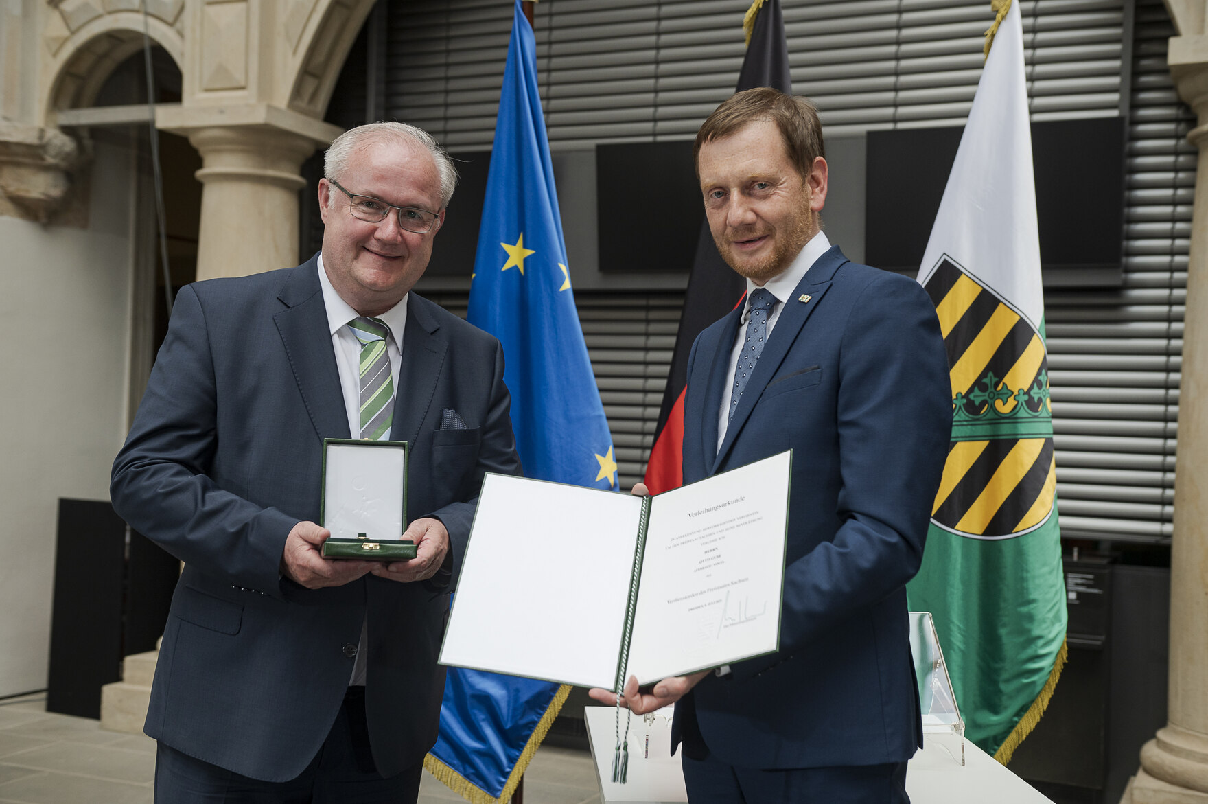 Ein Mann hält eine Urkunde in der Hand. Neben ihm steht ein weiterer Mann.