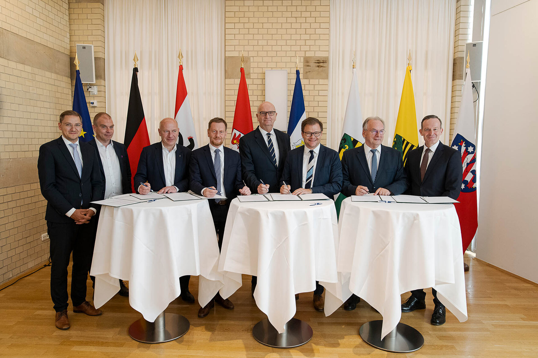 Männer und Frauen stehen an runden Konferenztischen. Hinter ihnen steht eine Fahnengalerie.