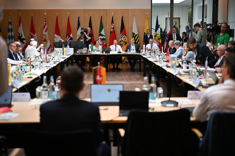 Männer und Frauen sitzen in einem Konferenzsaal umgeben von Flaggen der Bundesländer.